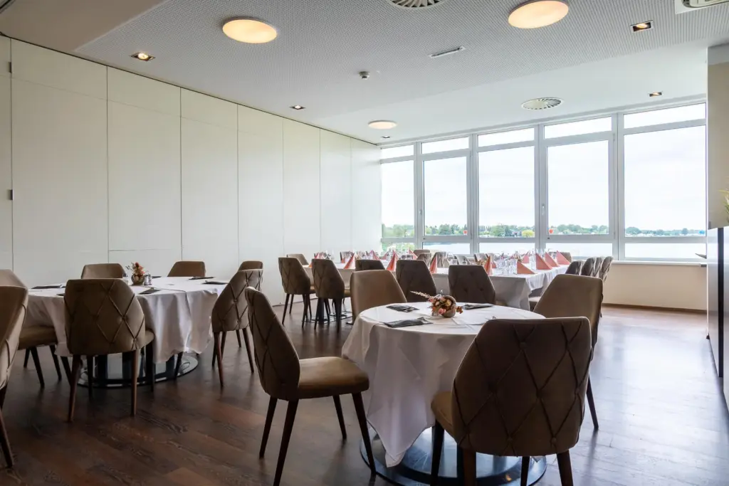 Ein moderner, heller Speisesaal mit runden, weiß gedeckten Tischen, braun gepolsterten Stühlen und großen Fenstern, die den Blick auf Bäume und Wasser freigeben. Der Raum ist leer und ordentlich eingerichtet.
