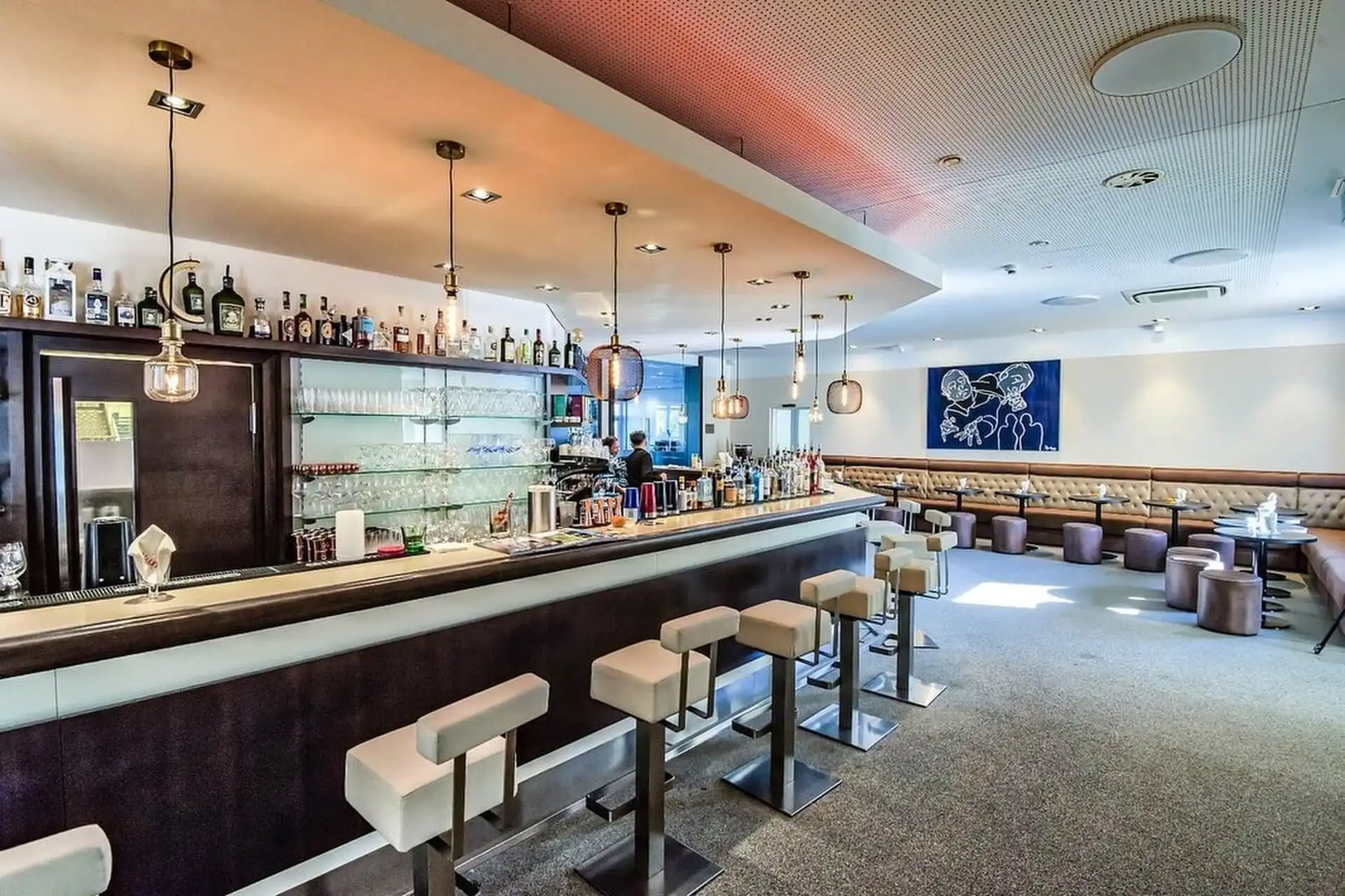 Moderne Bar mit hohen Hockern entlang einer Theke, Regalen mit Flaschen, Pendelleuchten über dem Kopf und einem Sitzbereich mit runden Tischen und gepolsterten Stühlen. Der Raum ist hell und modern mit einem blauen Wandkunstwerk.