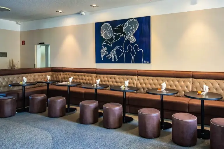 Ein modernes Wohnzimmer mit einer langen braunen getufteten Bank, runden braunen Hockern und kleinen schwarzen Tischen mit Servietten. Ein blaues abstraktes Kunstwerk mit zwei Figuren hängt an der beigen Wand über dem Sitzbereich.