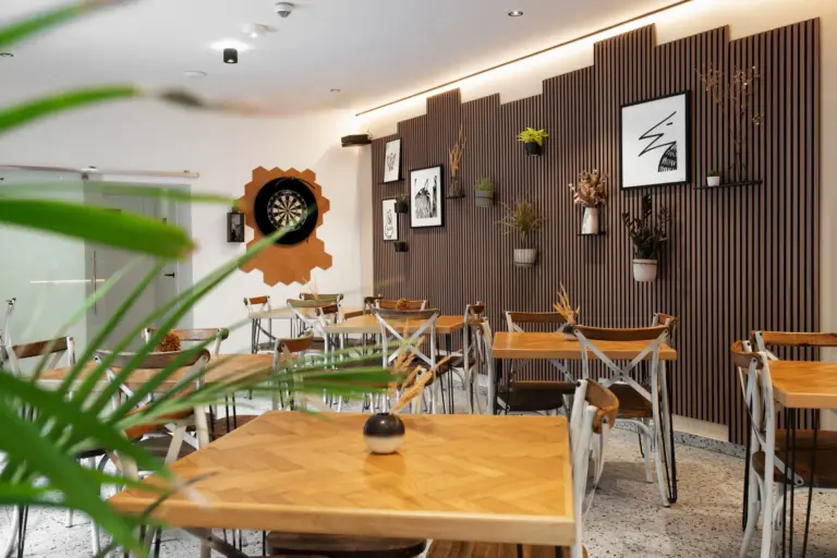 Ein modernes Café mit Holztischen und -stühlen, Pflanzen an den Wänden und gerahmter Kunst. Die Wände sind mit dekorativer Holzvertäfelung versehen, und in der Ecke steht eine Dartscheibe. Im Vordergrund ist eine Grünpflanze zu sehen.