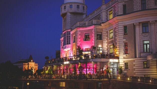 Ein großes, elegantes Gebäude mit einem Türmchen, das in der Abenddämmerung von rosa und violetten Lichtern angestrahlt wird, verfügt über eine belebte Außenterrasse, auf der Menschen essen und sich unterhalten.