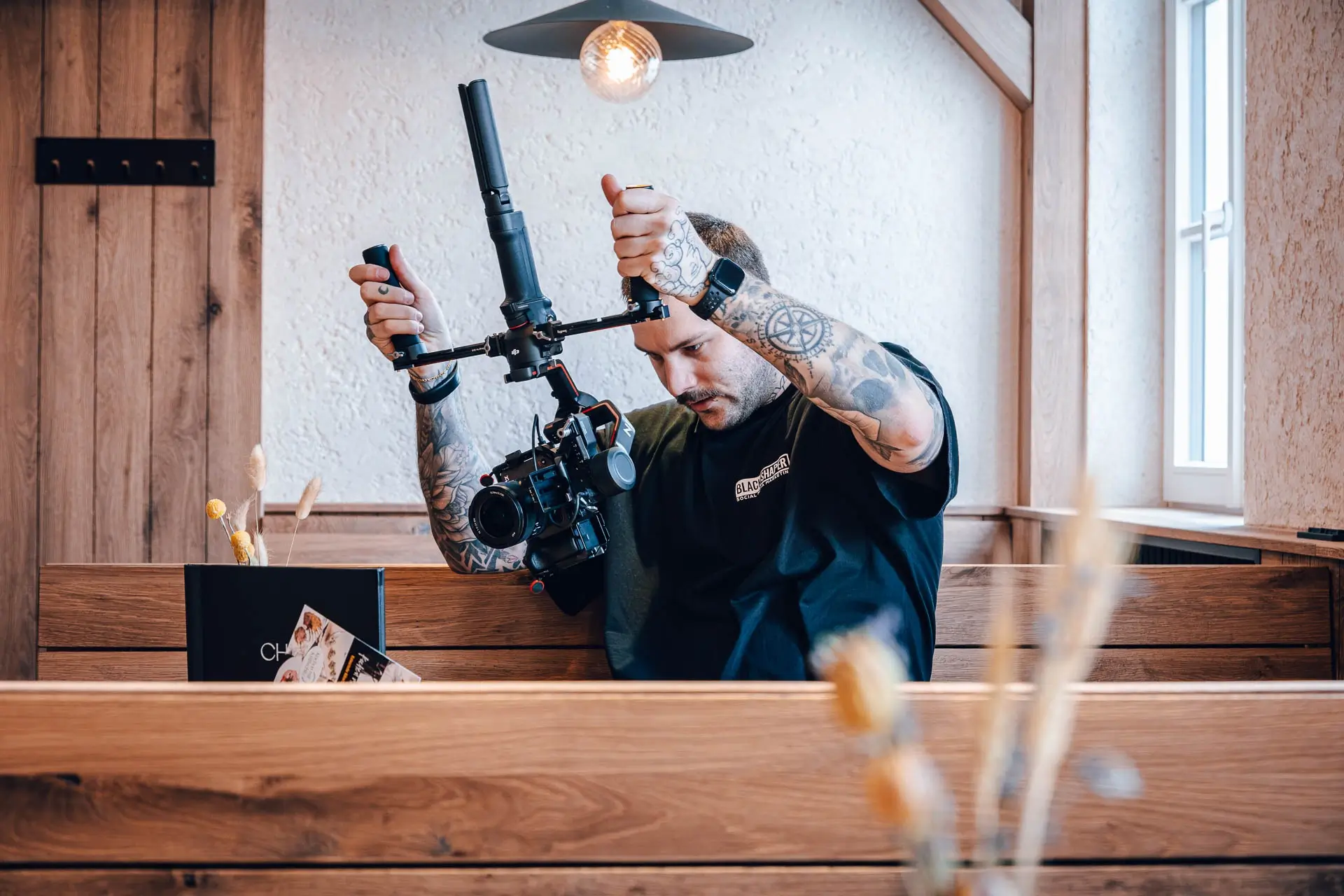 Eine tätowierte Person bedient eine professionelle Kameraausrüstung in einer Holzkabine und konzentriert sich auf die Aufnahmen. Das Sonnenlicht fällt durch ein nahe gelegenes Fenster und schafft eine gemütliche Innenatmosphäre.