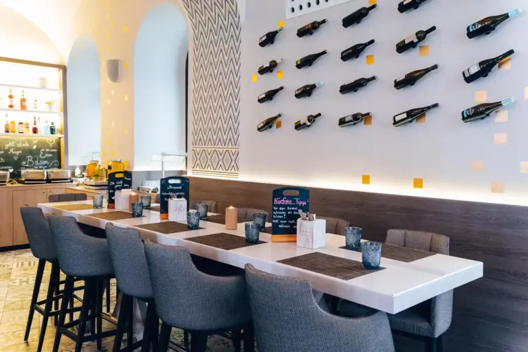 Modernes Restaurantinterieur mit einem langen Tisch und grauen Stühlen, an der Wand befestigten Weinflaschen als Dekoration und kleinen Kreidetafel-Menüs auf dem Tisch. Helle und luftige Atmosphäre mit stilvollen Designelementen.