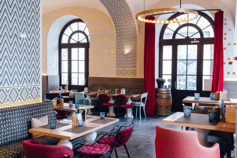 Ein gemütliches Restaurant mit gemusterten Wänden, gewölbten Fenstern, Holztischen, gemischten Stühlen, roten Vorhängen und einem runden Kronleuchter, die eine warme und einladende Atmosphäre schaffen.