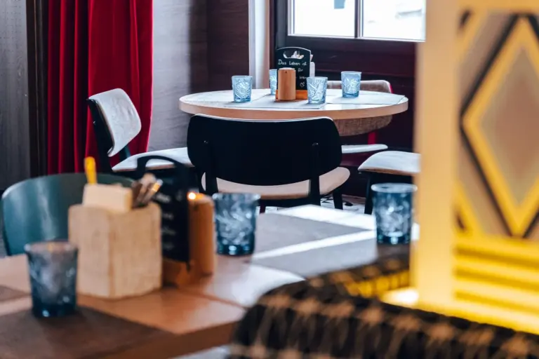Eine gemütliche Restaurantkulisse mit runden Tischen, schwarzen Stühlen und blau gemusterten Gläsern. Serviettenhalter und Gewürze stehen auf den Tischen, im Hintergrund sind rote Vorhänge und ein Fenster zu sehen.