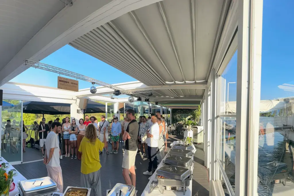 Eine Gruppe von Menschen steht in einem Halbkreis und hört einem Redner auf einer überdachten Außenterrasse des Vienna City Beach Club zu, auf der Tische mit Buffet und Essenstabletts stehen. Große Fenster bieten an einem sonnigen Tag einen Blick auf Wasser und Grünflächen.