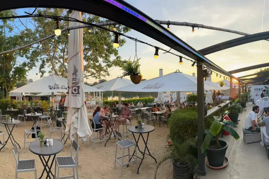 Im Vienna City Beach Club sitzen die Leute an Tischen im Freien auf einer Sandterrasse unter großen weißen Sonnenschirmen und entspannen sich. Lichterketten und Pflanzen schmücken den Bereich, während die Sonne im Hintergrund untergeht und eine perfekte Strandcafé-Atmosphäre schafft.