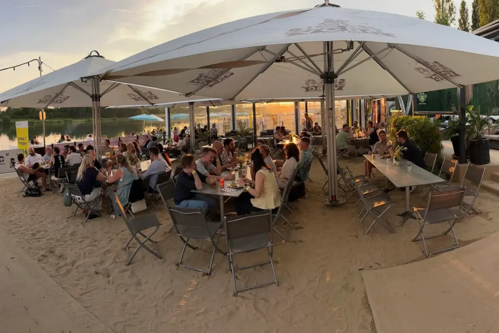 Im Vienna City Beach Club sitzen die Menschen bei Sonnenuntergang an Tischen unter großen Sonnenschirmen auf einer sandigen Terrasse in der Nähe eines Sees und genießen das Essen und die Gespräche in einer entspannten, strandähnlichen Umgebung.