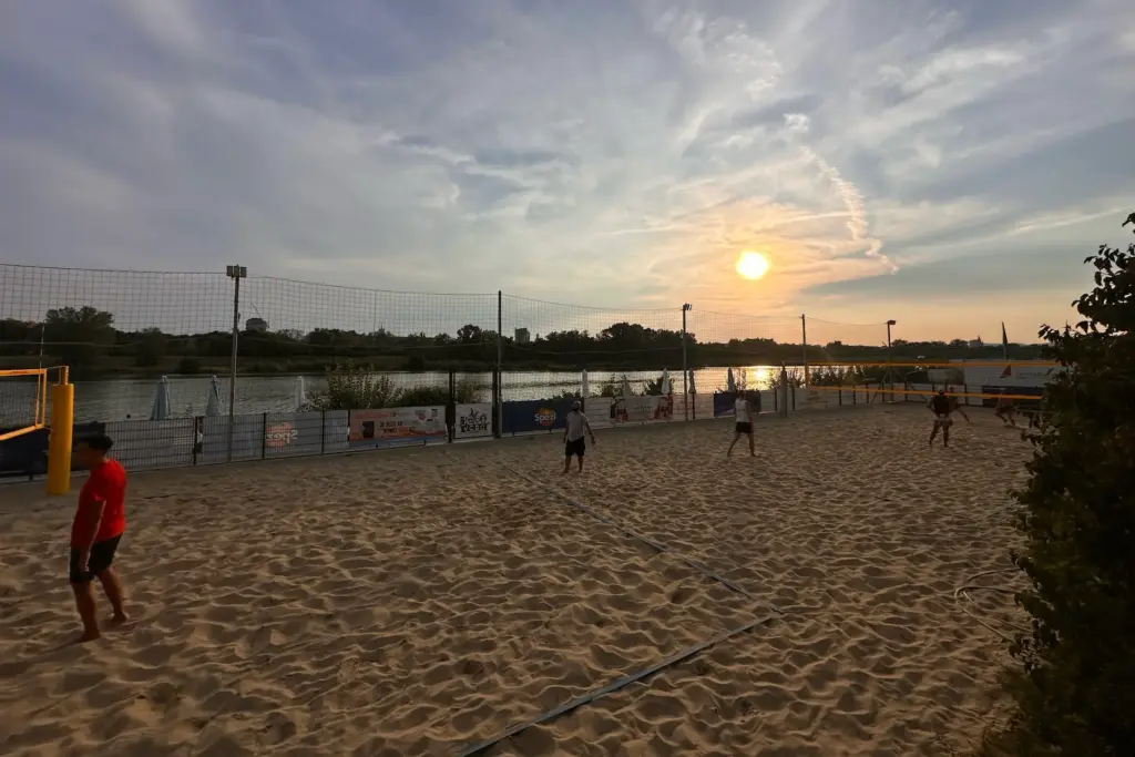 Menschen spielen Beachvolleyball auf einem Sandplatz in der Nähe eines Gewässers bei Sonnenuntergang. Der Himmel ist teilweise bewölkt, und Bäume säumen das gegenüberliegende Ufer. Die Szene strahlt eine entspannte Atmosphäre im Freien aus.