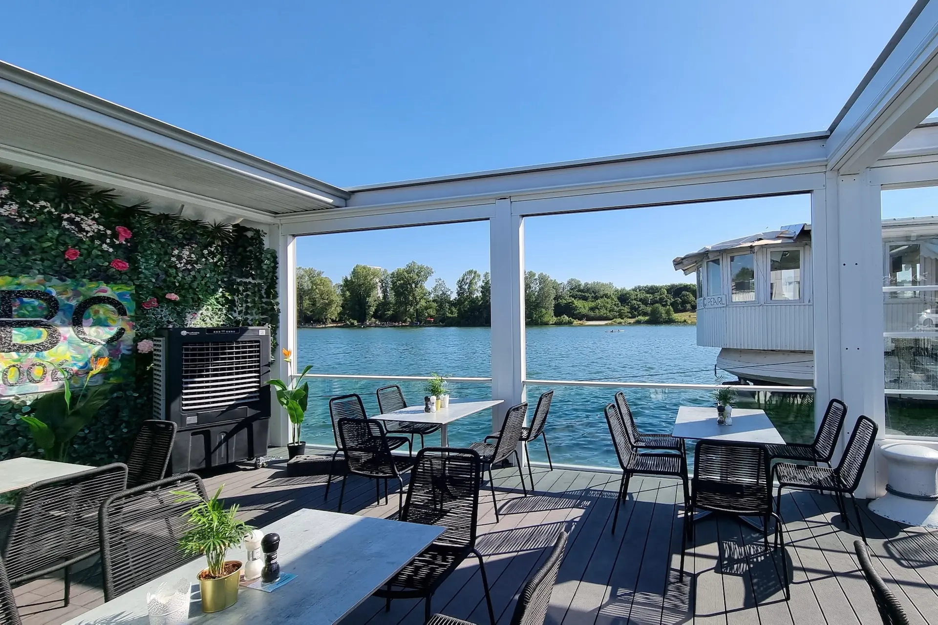 Terrasse des Außenrestaurants mit schwarzen Stühlen und Tischen, mit Blick auf einen ruhigen Fluss. Topfpflanzen schmücken den Bereich, und die Aussicht umfasst grüne Bäume und einen blauen Himmel unter hellem Sonnenlicht.