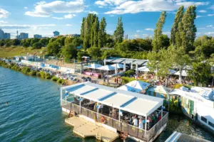 Vienna City Beach Club Ein lebhaftes Restaurant am Flussufer mit Sitzplätzen im Freien und weißen Sonnenschirmen, umgeben von Bäumen und Menschen, die den sonnigen Tag genießen. Im Hintergrund sind die Skyline der Stadt und der klare blaue Himmel zu sehen.