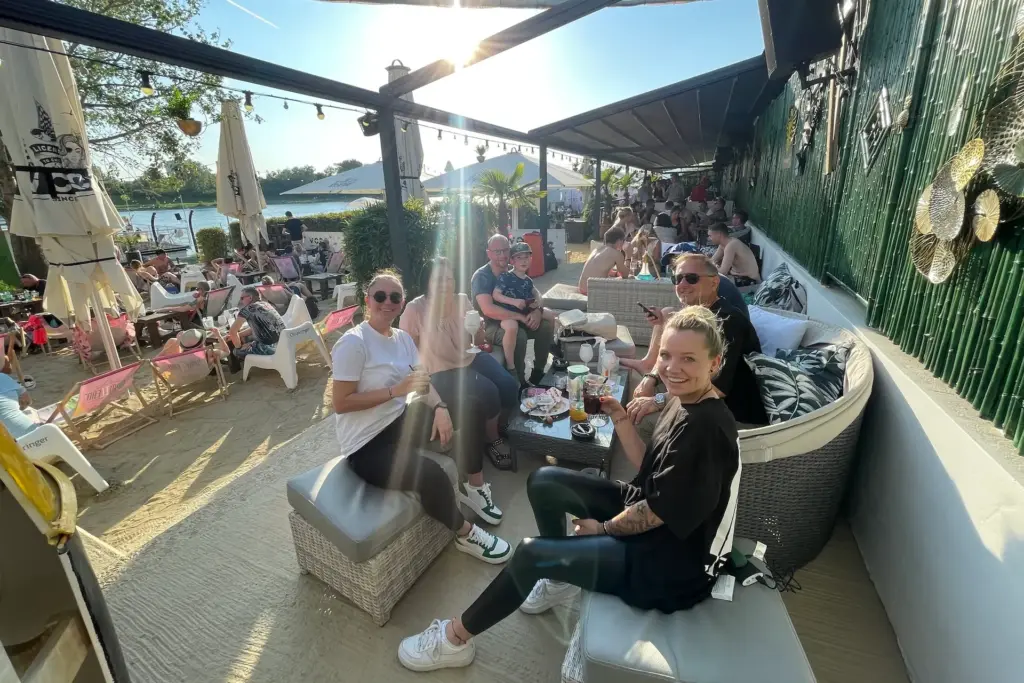 Eine Gruppe von Menschen entspannt sich und lächelt an einer Strandbar im Freien. Sie sitzen auf gepolsterten Stühlen um einen niedrigen Tisch mit Getränken und Snacks, während andere Gäste sich im sonnigen Hintergrund unterhalten.