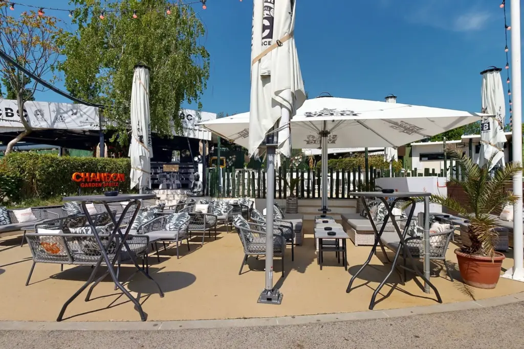 Der Café-Bereich im Freien mit weißen Stühlen, Tischen und großen Sonnenschirmen an einem sonnigen Tag; Topfpflanzen und Lichterketten vermitteln die entspannte Atmosphäre des Vienna City Beach Club.
