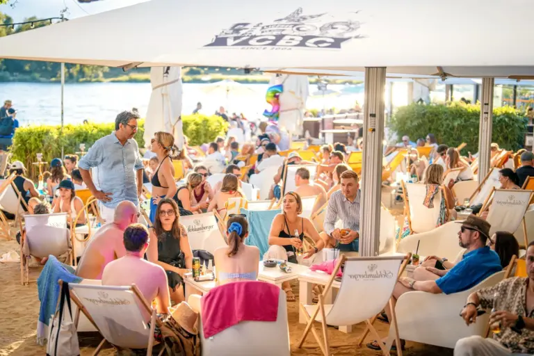 Eine lebhafte Strandbarszene mit vielen Menschen, die sich auf Liegestühlen unter einem großen Sonnenschirm entspannen, Kontakte knüpfen und an einem sonnigen Tag in der Nähe des Ufers Getränke genießen.