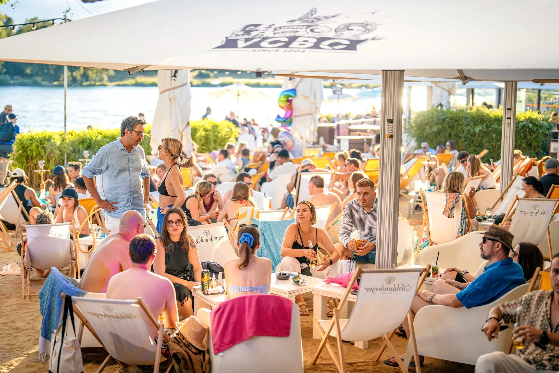 Eine lebhafte Strandbarszene mit vielen Menschen, die sich auf Liegestühlen unter einem großen Sonnenschirm entspannen, Kontakte knüpfen und an einem sonnigen Tag in der Nähe des Ufers Getränke genießen.