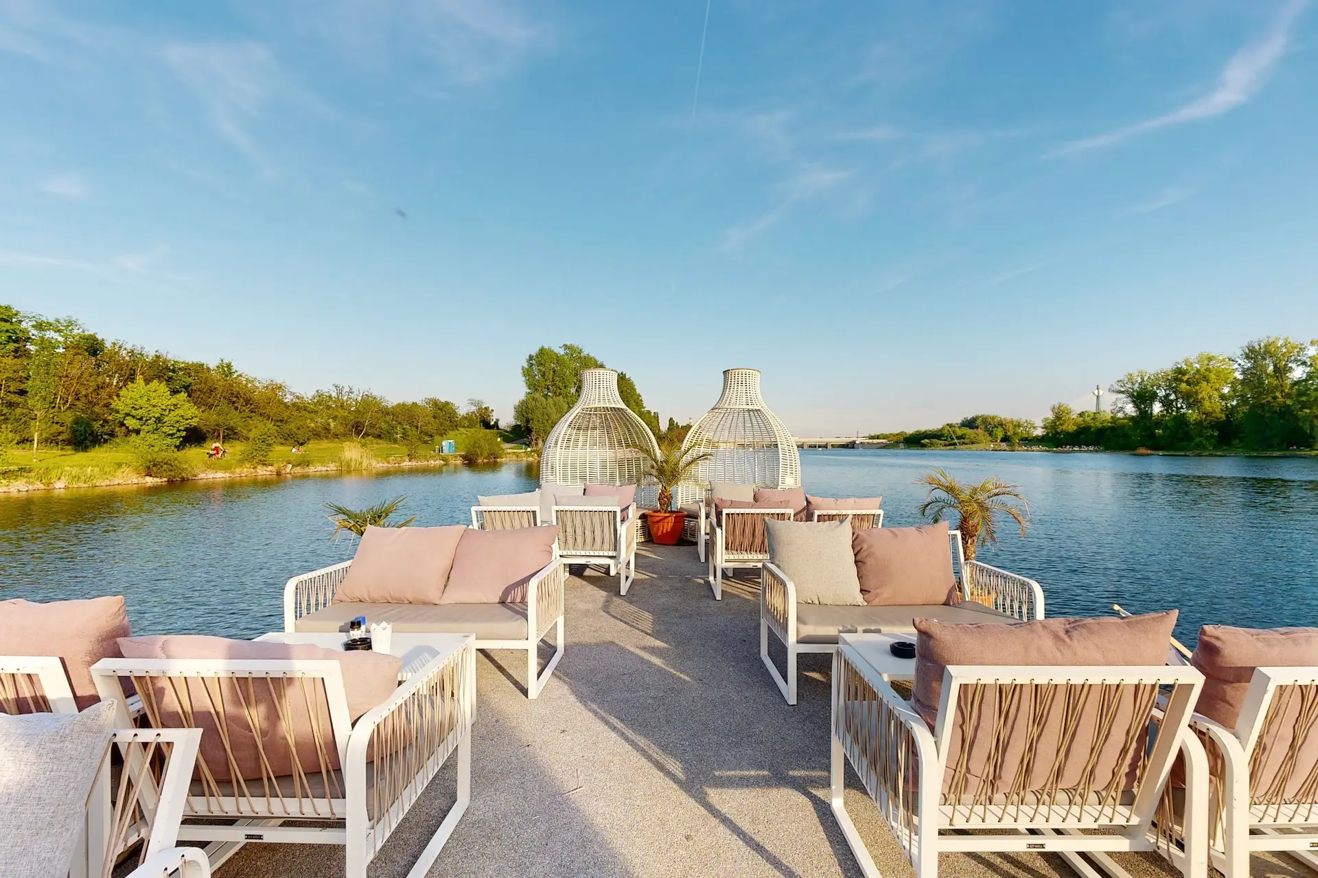 Lounge-Bereich im Freien mit gepolsterten Stühlen und Tischen auf einem Steg über einem ruhigen Fluss, umgeben von Grün, mit zwei dekorativen Weidenstrukturen und klarem blauen Himmel.