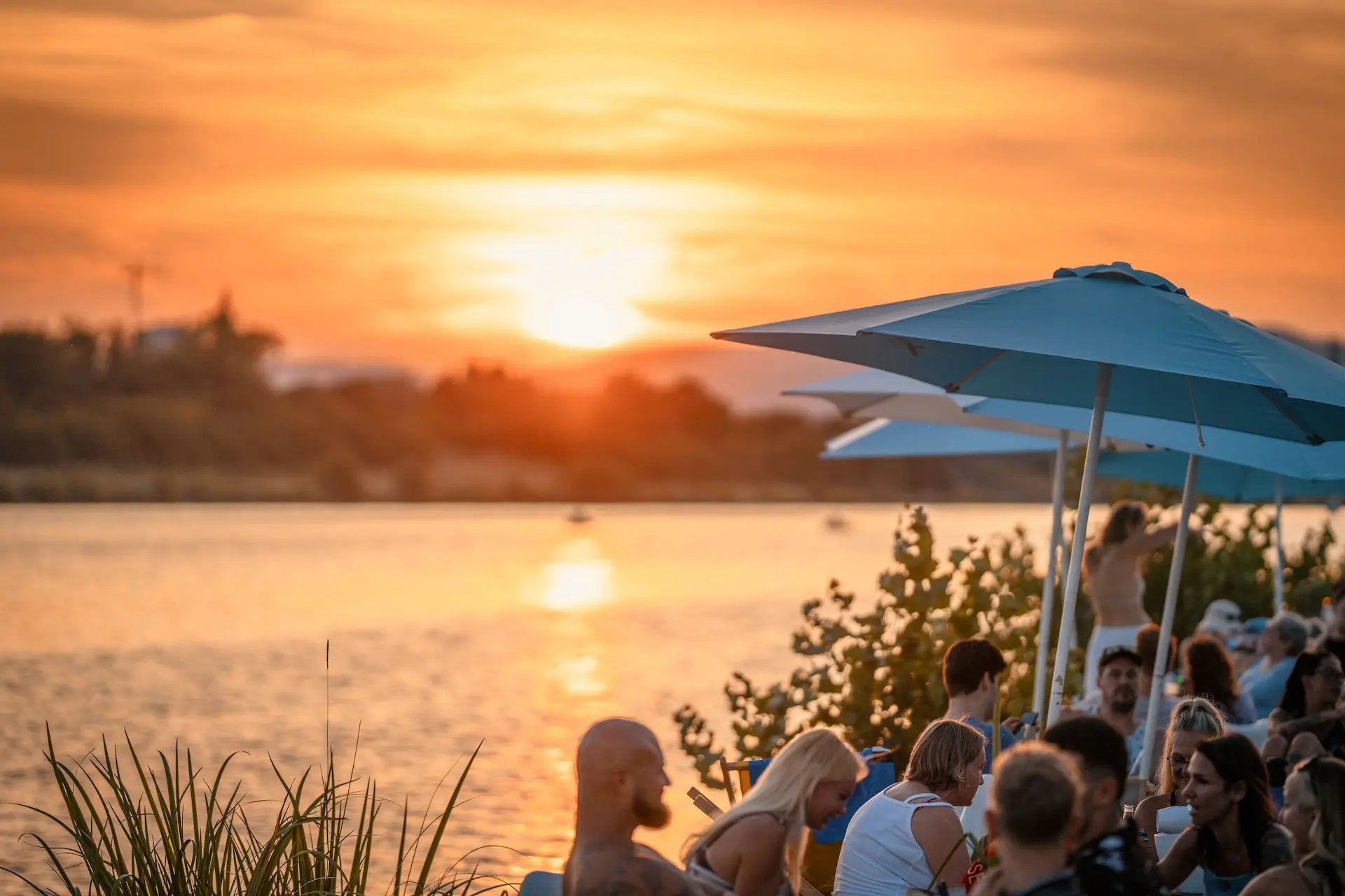 Menschen versammeln sich bei Sonnenuntergang am Flussufer und sitzen unter blauen Sonnenschirmen. Der Himmel ist orange, und die Sonne leuchtet über dem Wasser und schafft eine warme, entspannte Atmosphäre.