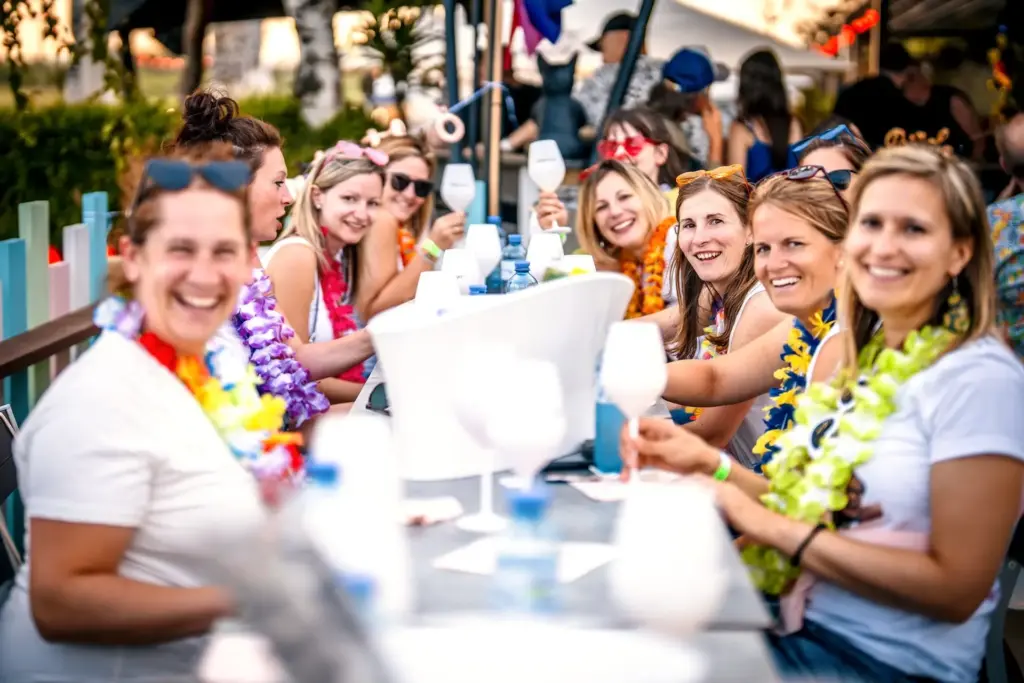 Eine Gruppe lächelnder Frauen mit bunten Blumenkränzen sitzt an einem langen Tisch im Freien, genießt Getränke und feiert gemeinsam in festlicher Atmosphäre.