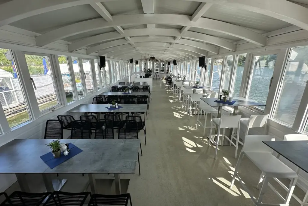 Ein heller, moderner Essbereich auf einem Boot mit großen Fenstern, grauen Tischen, schwarzen und weißen Stühlen, blauen Tischsets und kleinen Topfpflanzen, mit Blick auf das Wasser und die Grünanlagen draußen.