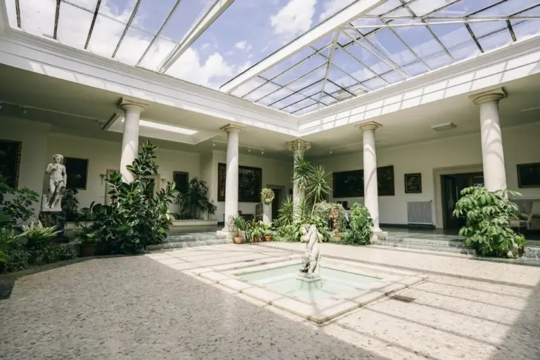 Ein heller Innenhof mit einer Glasdecke, klassischen Statuen, Topfpflanzen, Säulen und gerahmten Kunstwerken an den Wänden; Sonnenlicht strömt herein und beleuchtet einen kleinen Brunnen und eine Statue in der Mitte.