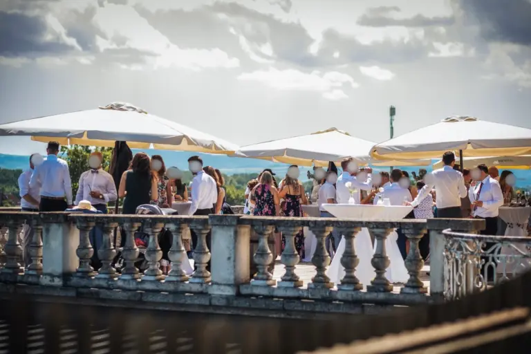 Eine Gruppe von Menschen versammelt sich im Freien unter großen weißen Schirmen zu einem gesellschaftlichen Ereignis. Sie stehen um Cocktailtische auf einer Terrasse mit dekorativem Geländer, unter einem teilweise bewölkten Himmel.