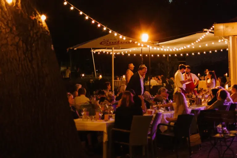 Abends speisen die Gäste im Freien unter Lichterketten; das Servicepersonal serviert das Essen, und die Gäste sitzen in einer warmen, festlichen Atmosphäre an den Tischen.