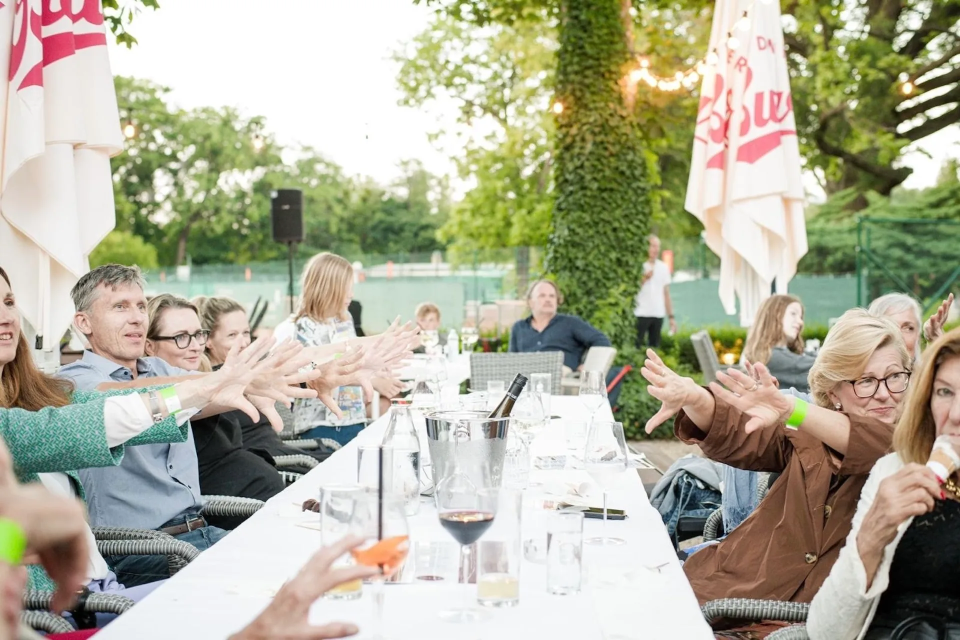 Menschen sitzen an einem langen Tisch im Freien, strecken ihre Hände in Richtung Kamera aus; im Hintergrund sind Bäume, Sonnenschirme und Getränke auf dem Tisch zu sehen. Die Atmosphäre wirkt lebhaft und gesellig.