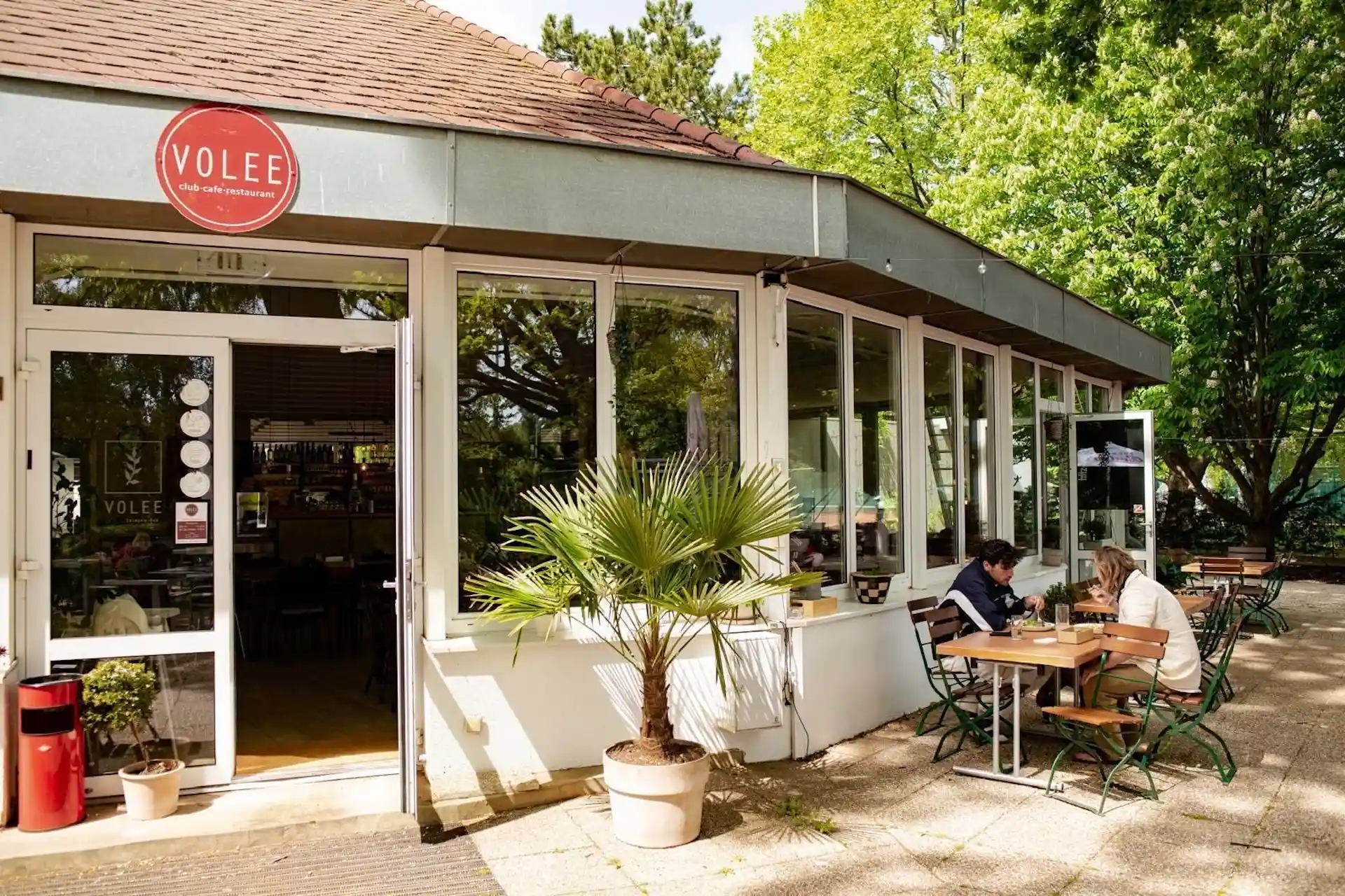Sitzgelegenheiten im Außenbereich eines Cafés mit großen Fenstern, einem roten Schild mit der Aufschrift „VOLEE“ und Menschen, die an einem Tisch sitzen. Im Hintergrund sind grüne Bäume und eine Topfpalme zu sehen, und Sonnenlicht fällt auf die Szene.