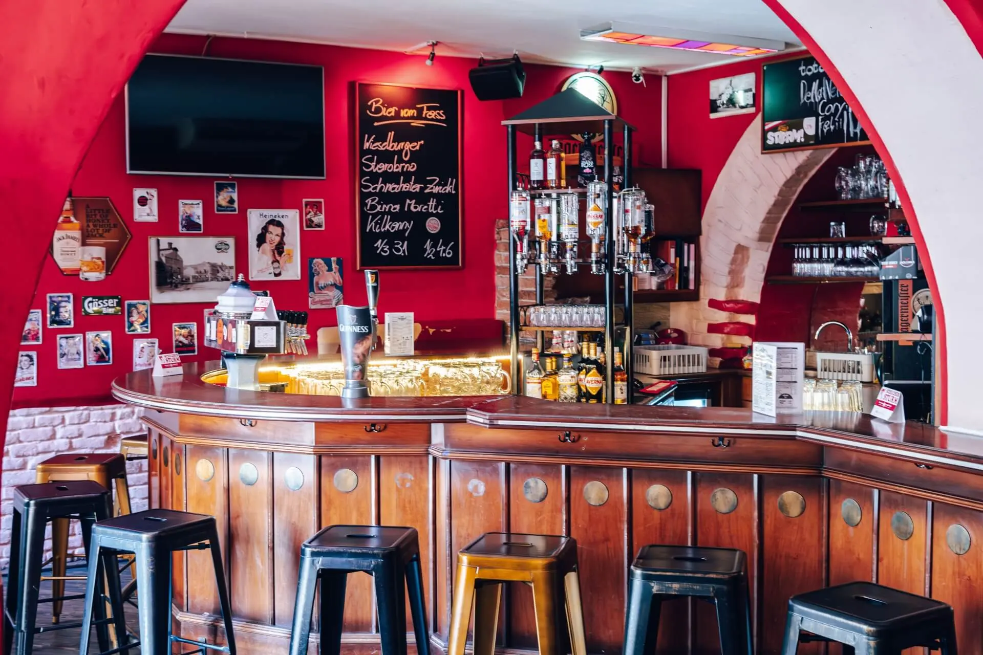 Gemütliche Bar mit einem geschwungenen Holztresen, Hockern, Flaschenregalen, einer Speisekarte aus Kreide und mit Fotos und Postern dekorierten Wänden. Der Raum hat hellrote Wände und eine entspannte Atmosphäre.