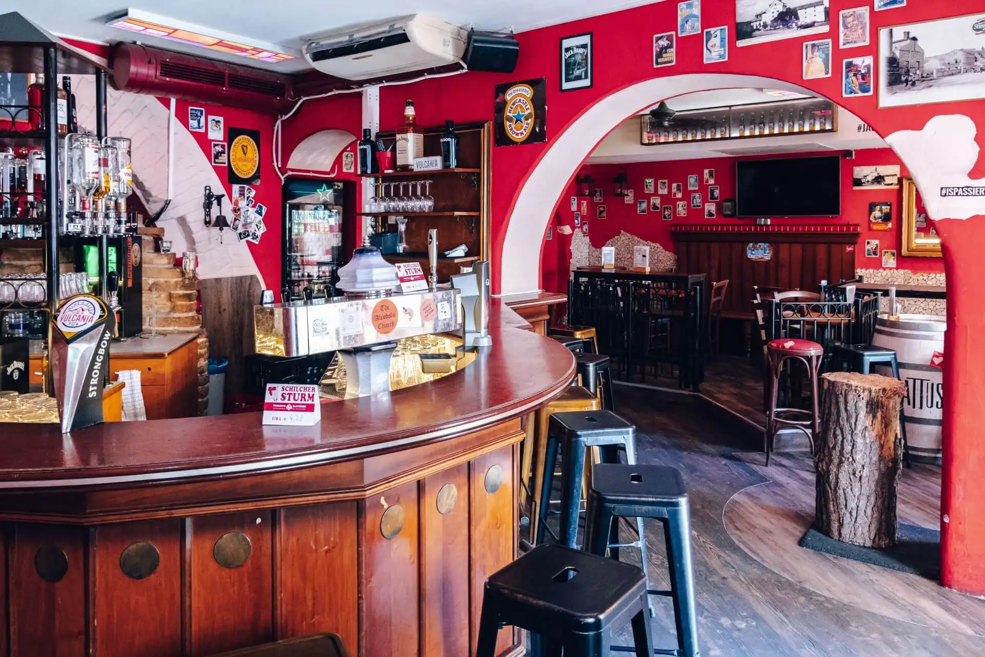 Eine gemütliche Bar mit einem geschwungenen Holztresen, schwarzen Hockern, roten Wänden mit gerahmten Fotos, Regalen mit Flaschen und einem kleinen Essbereich mit Tischen und Stühlen im Hintergrund.