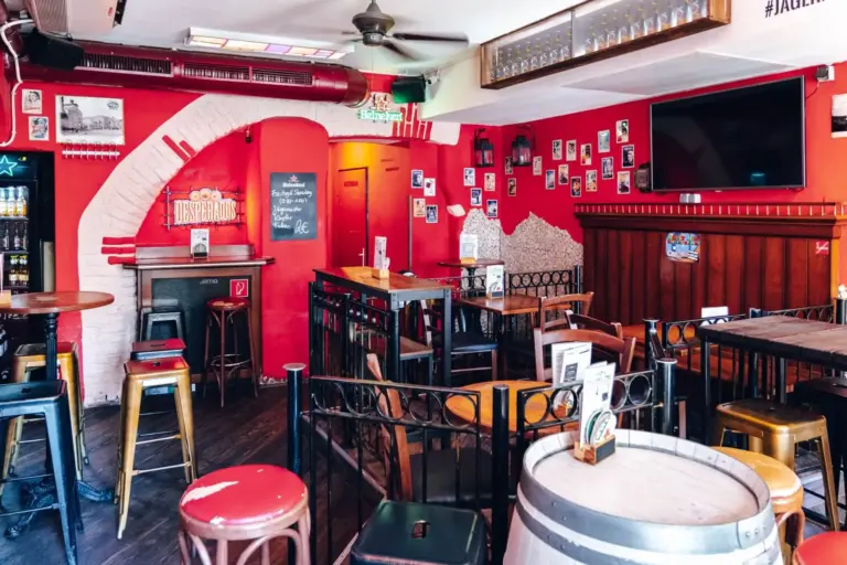 Eine gemütliche Bar mit roten Wänden, Holztischen und -hockern, einer Speisekarte auf Kreidetafeln, Wandfotos und einem kleinen Bühnenbereich. Der Raum ist leer, das Sonnenlicht strömt herein und dekorative Fässer dienen als Tische.