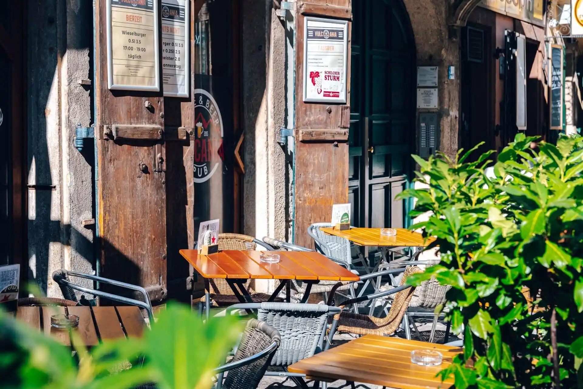 Außencafé mit Holztischen und Korbstühlen vor rustikalen Holztüren und Steinwänden. Die Speisekarten hängen an der Wand, und Grünpflanzen umrahmen teilweise den Sitzbereich und schaffen eine einladende, sonnige Atmosphäre.