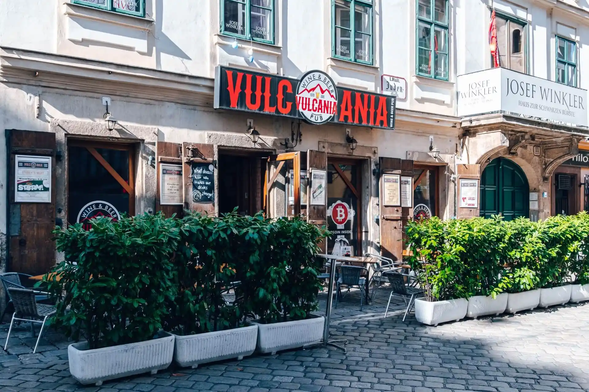 Sitzbereich im Freien mit Tischen und Stühlen vor dem Restaurant Vulcania auf einer Kopfsteinpflasterstraße, mit Grünpflanzen in weißen Pflanzkübeln, die den Innenhof vom Gehweg trennen.
