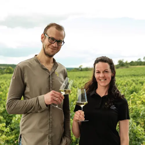 Ein Mann und eine Frau stehen in einem Weinberg, lächeln und halten Gläser mit Weißwein in der Hand. Im Hintergrund sind grüne Weinreben und ein bewölkter Himmel zu sehen.