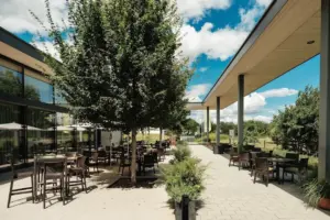 Die Außenterrasse eines modernen Restaurants mit Tischen und Stühlen unter Bäumen und einem überdachten Gehweg, umgeben von Grün, an einem sonnigen Tag mit blauem Himmel und vereinzelten Wolken.