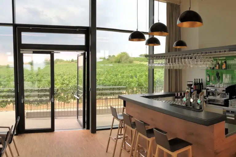 Moderne Bar mit Holzhockern und Hängelampen, großen Glasfenstern mit Blick auf einen grünen Weinberg unter einem wolkenverhangenen Himmel und Regalen mit Flaschen und Gläsern hinter dem Tresen.