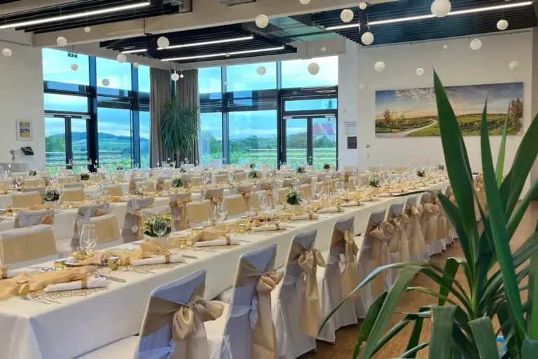 Eleganter Bankettsaal mit langen, weiß gedeckten Tischen, goldenen Schleifen an den Stühlen, ordentlich gedeckten Tischen und großen Fenstern, die den Blick auf eine malerische Landschaft freigeben. Weiße Hängelampen tragen zur festlichen Atmosphäre bei.