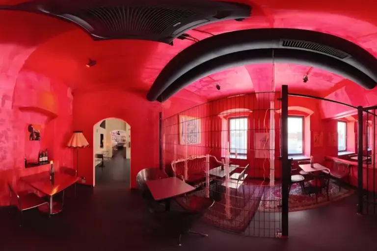 Ein gemütliches, modernes Café mit leuchtend roten Wänden und Decken, schwarzen Akzenten, Tischen und Stühlen, einer Trennwand aus Draht, großen Fenstern, einem Teppich und einer Stehlampe, die eine eklektische und künstlerische Atmosphäre schaffen.