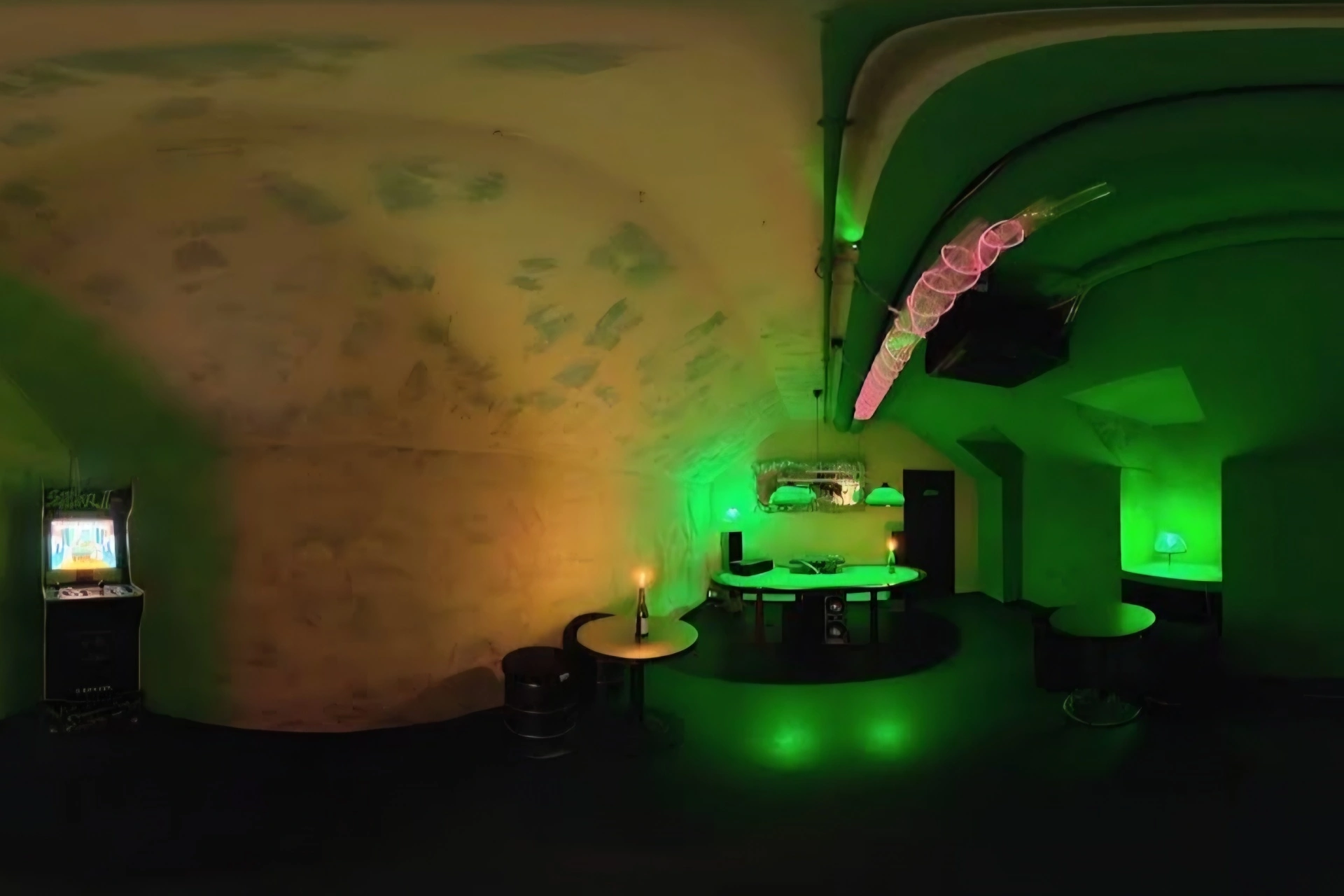 Ein schwach beleuchteter Raum mit gewölbten Decken, grüner Umgebungsbeleuchtung, Tischen, einem Bartresen, einer Lampe und einem Spielautomaten in der Ecke. Rosa Lichter hängen von der Decke und schaffen eine stimmungsvolle, futuristische Atmosphäre.