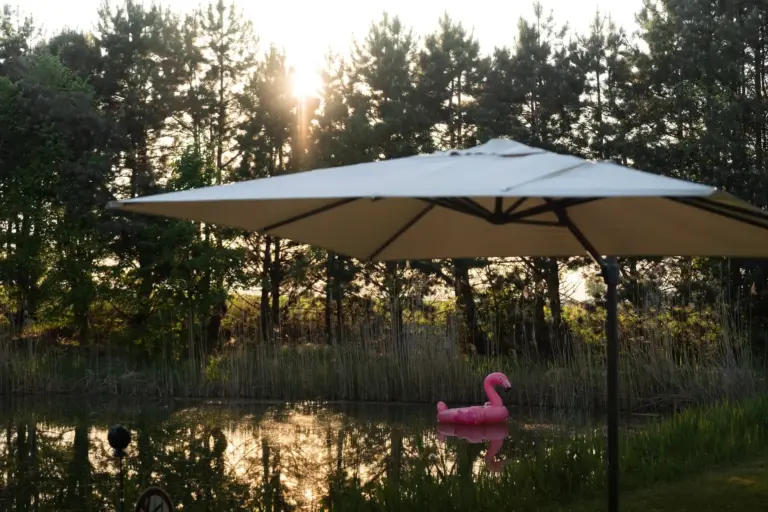 Ein großer weißer Terrassenschirm beschattet einen Bereich neben einem Teich, in dem ein rosa aufblasbarer Flamingo auf dem Wasser schwimmt. Im Hintergrund sind hohes Gras und Bäume zu sehen, durch die das Sonnenlicht scheint.