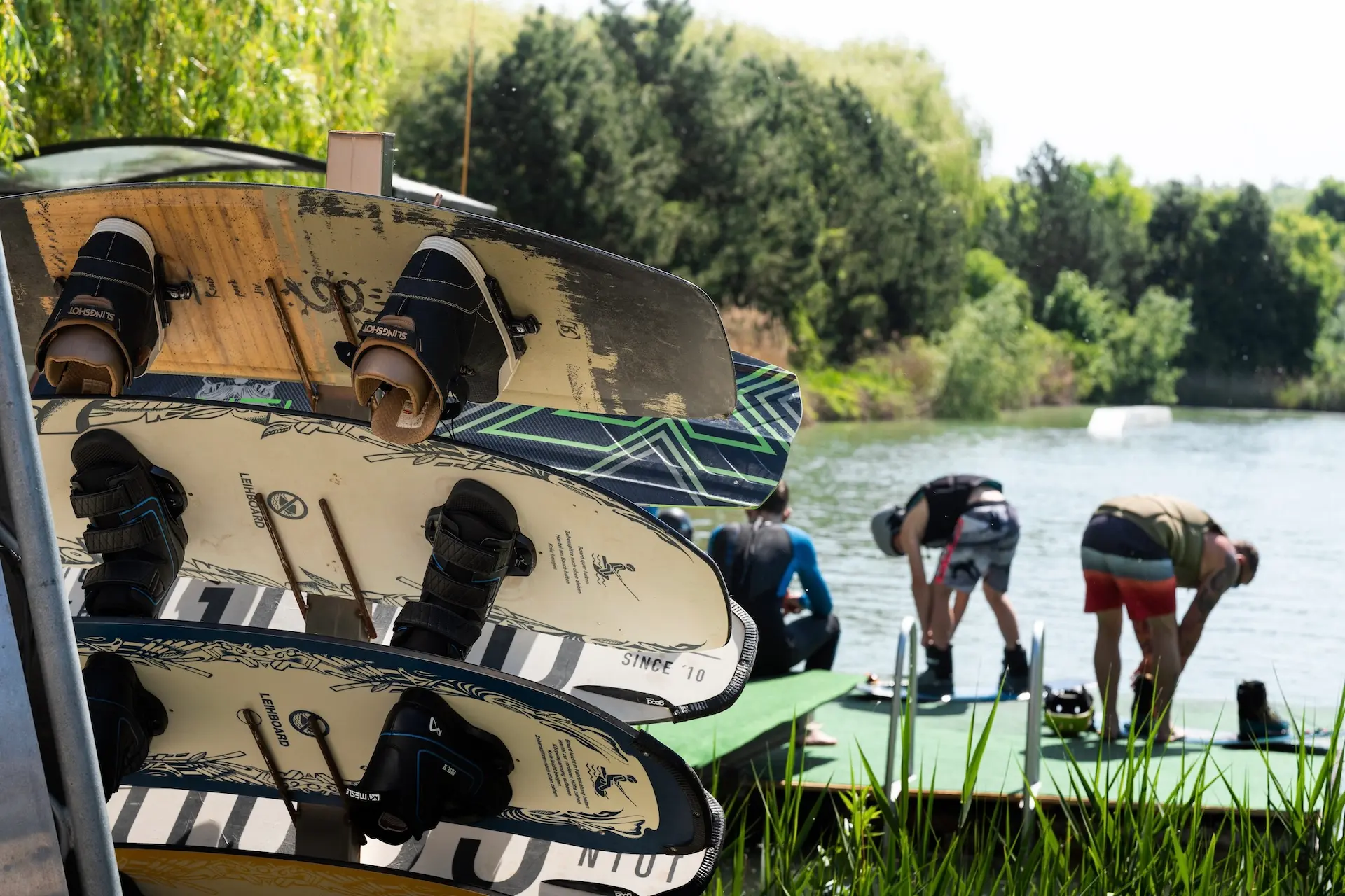 Im Vordergrund sind mehrere Wakeboards gestapelt, während sich Menschen in Badeanzügen an einem Steg an einem See vorbereiten, umgeben von grünen Bäumen und Pflanzen an einem sonnigen Tag.