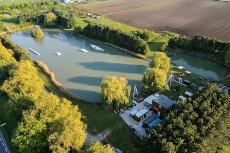 Wakeground Luftaufnahme eines von Bäumen und Feldern umgebenen Sees mit Rampen und Seilen für Wakeboarding und mehreren Gebäuden und Geräten am Ufer.