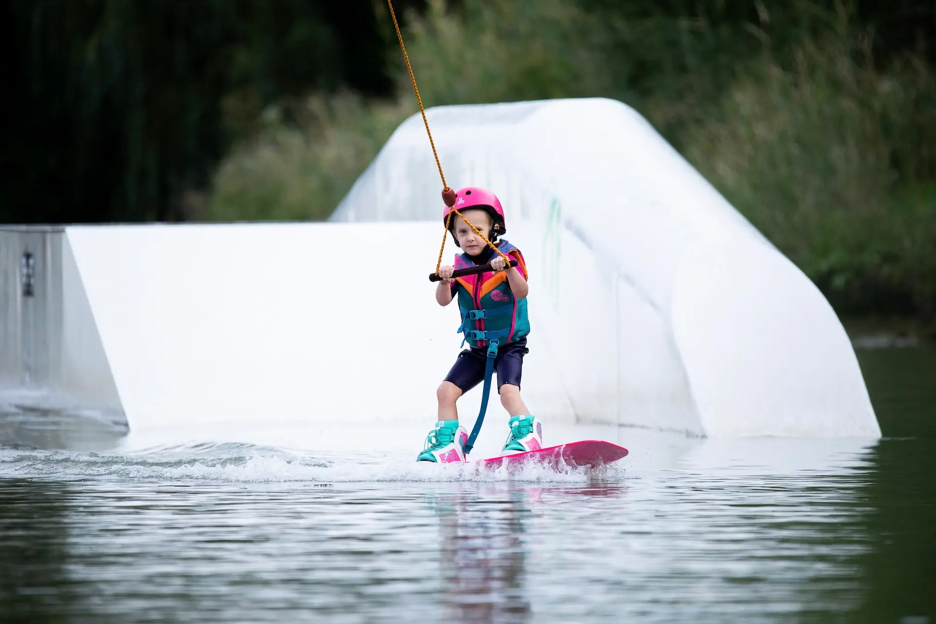 Ein kleines Kind, das einen rosa Helm und eine Schwimmweste trägt, fährt auf einem See Wakeboard und hält sich an einem Seil fest. Das Kind lächelt und balanciert auf einem rosa Brett, mit einer weißen Rampe und grünen Bäumen im Hintergrund.