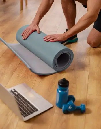 Auf einem Holzboden kniende Person, die eine Yogamatte zusammenrollt - bereit, den Weihnachtsspeck loszuwerden. Im Vordergrund sind ein Laptop, eine blaue Wasserflasche und zwei blaue Hanteln zu sehen.