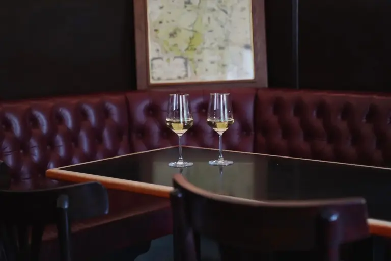 Zwei Gläser Weißwein stehen auf einem schwarzen Tisch in einer gemütlichen Kabine mit kastanienbraunen Ledersitzen; an der dunklen Wand hinter ihnen hängt eine gerahmte Landkarte.