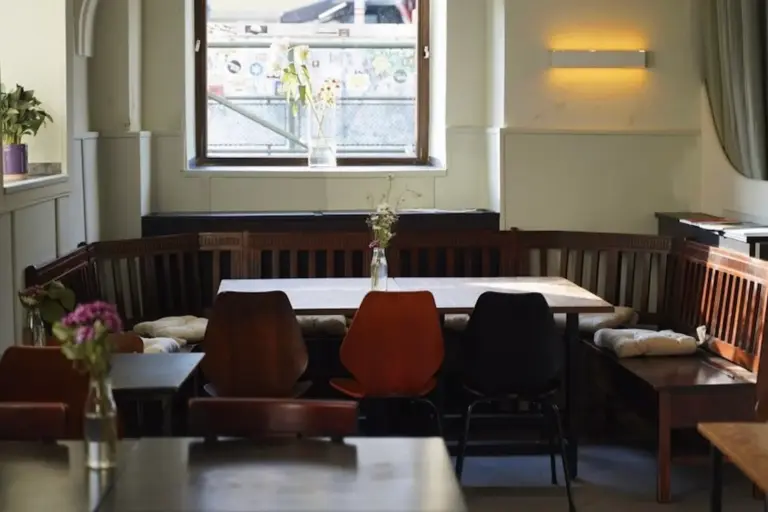 Ein gemütliches Café mit Holzbänken und -stühlen um Tische herum, Licht, das durch ein Fenster fällt, und Vasen mit Blumen als Tafelaufsatz schaffen eine warme, einladende Atmosphäre.