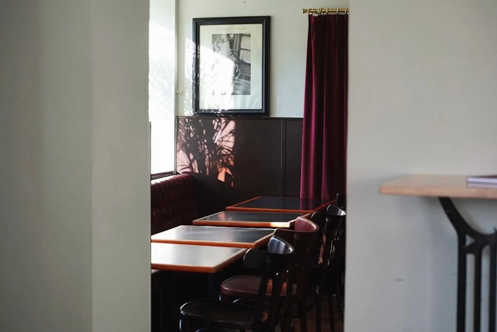 Ein kleines, sonnendurchflutetes Eckcafé mit leeren schwarzen Stühlen, Holztischen, einer kastanienbraunen Sitzecke und einem weinroten Vorhang. An der Wand über dem Tisch hängt ein gerahmtes Schwarz-Weiß-Foto.