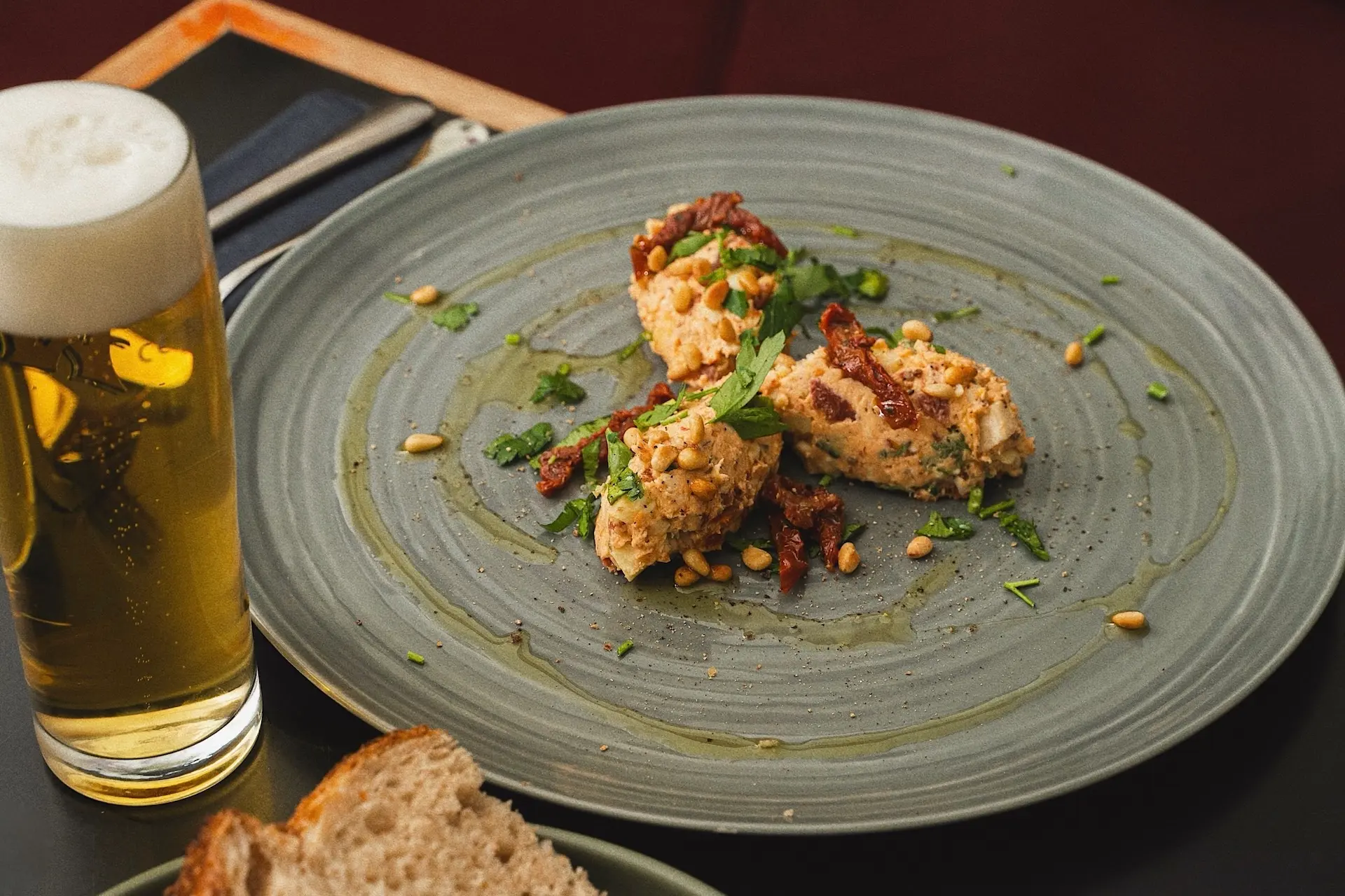 Ein grauer Teller mit drei Kugeln Aufstrich, garniert mit Kräutern, sonnengetrockneten Tomaten und Pinienkernen, neben einem Glas Bier und Brotscheiben auf einem dunklen Tisch.