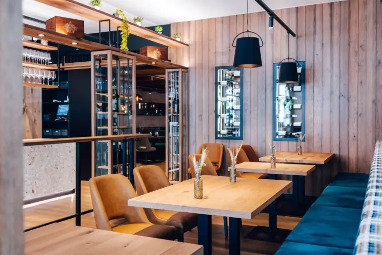 Ein modernes Restaurant mit Holztischen, hellbraunen Stühlen, blauen Sitzkissen, schwarzen Hängelampen, Glastrennwänden und hölzernen, mit Pflanzen und Spiegeln dekorierten Wandpaneelen.