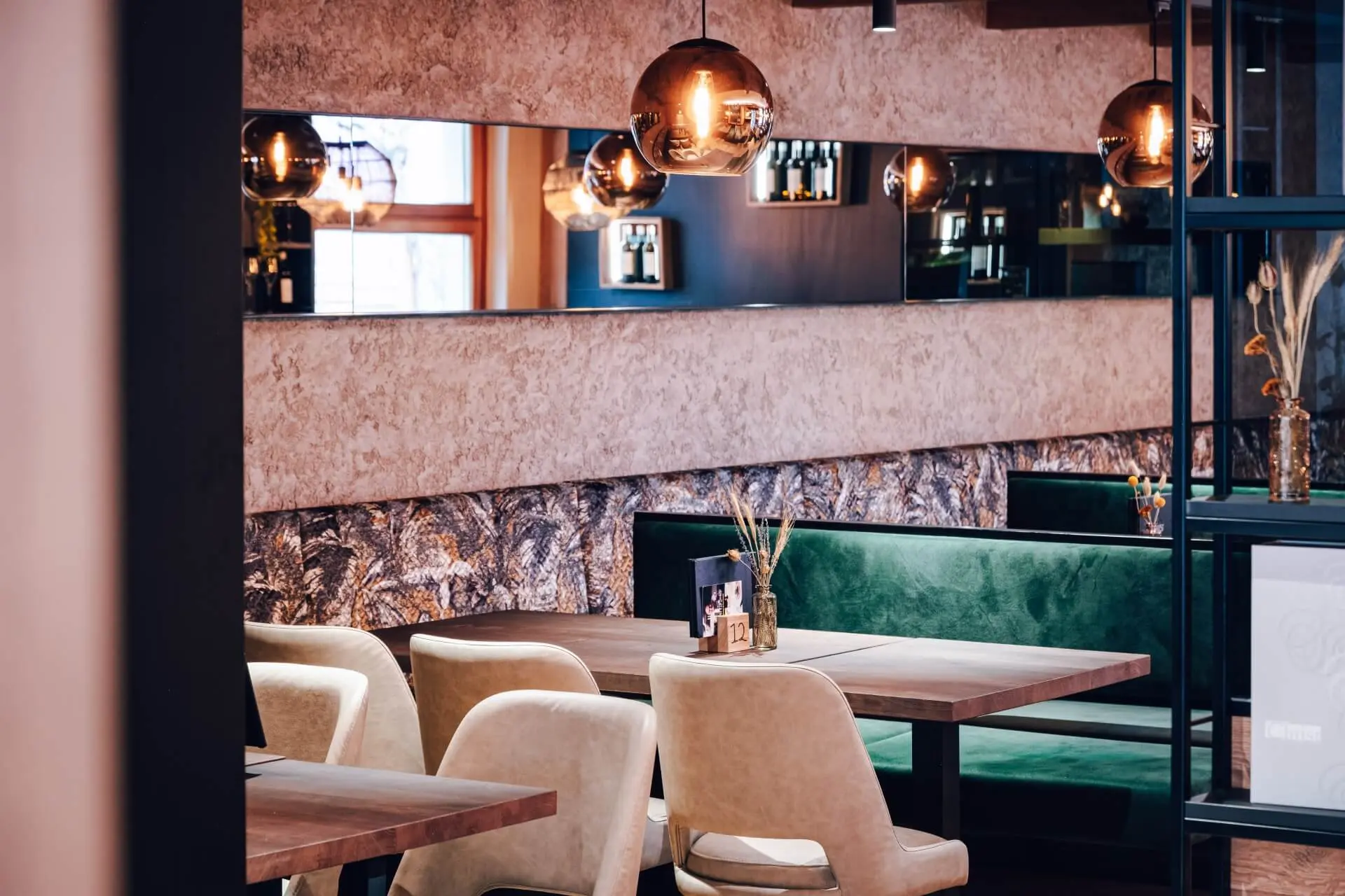 Ein modernes Restaurant mit cremefarbenen Plüschsesseln, grünen Samtbänken, Holztischen, hängenden Pendelleuchten und einem großen Spiegel, der die gemütliche, stilvolle Einrichtung widerspiegelt.